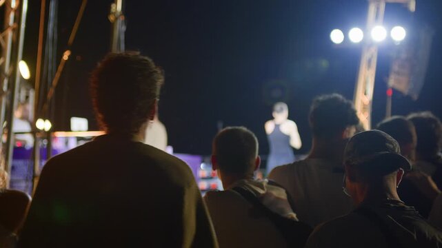Rear view of young man cheering in a lively concert crowd at night. Flashing stage lights and artist performance capture fan energy and immersive live music vibe.