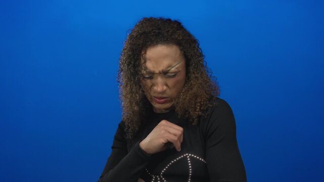 Woman with curly hair and black top coughs in blue studio setting with furrowed brows and clenched fist near chest; disgust.
