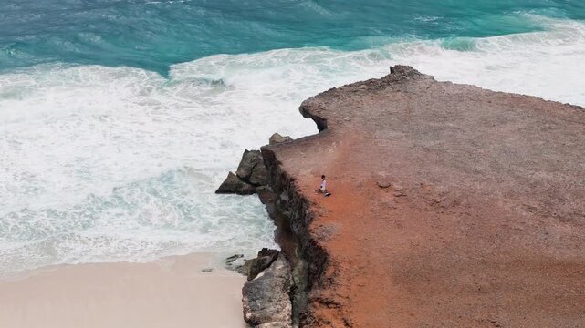 Toma a&eacute;rea de dron de una pareja de j&oacute;venes admirando y explorando el paisaje, sobre un acantilado rocoso y al fondo el mar azul con olas impactando en la costa de Aruba. &Eacute;pico e inspirador.