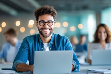 A smiling young man with glasses works on a laptop in a bright, modern office with colleagues blurred in the background.