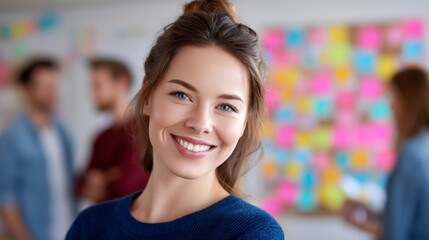 A cheerful young woman smiles confidently with blurred colleagues and colorful sticky notes in the background, suggesting a creative office environment.