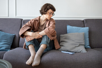 Thoughtful woman sadly looking on phone sits on couch waiting call or message feeling loneliness,...