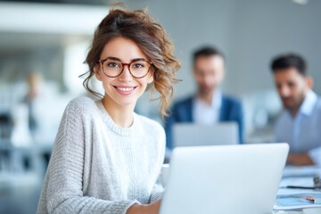 A young woman with glasses smiles confidently while working on a laptop in a modern office, with two colleagues blurred in the background.