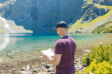 Male traveler standing near a scenic alpine lake with a map in hand Admiring the mountain view under clear skies Navigation and adventure in nature
