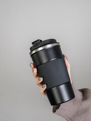 Female hand in brown sweater holding black thermos mug with coffee on grey background
