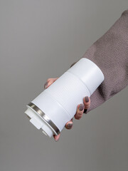 Female hand in brown sweater holding white thermos mug with coffee on grey background
