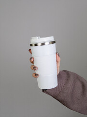 Female hand in brown sweater holding white thermos mug with coffee on grey background
