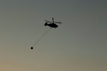 A firefighting helicopter carrying water to extinguish a pine forest fire in the evening of a hot...