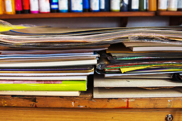 A colorful stack of sketchbooks, drawing pads, paper portfolios,  layers of paper, canvas boards, paint tubes, and mixed media folders on a wooden desk in an art studio.
