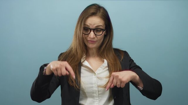 Woman pointing finger downward in blue studio wearing glasses and blazer with white shirt and thoughtful frown; doubt.