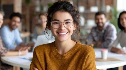 A confident young woman wearing glasses and a mustard sweater smiles warmly in a casual office setting with colleagues blurred in the background.