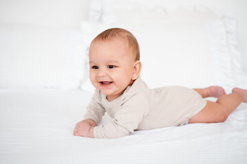 a baby, a smiling baby on a white bed on his tummy at home, a healthy, joyful little baby boy in a...