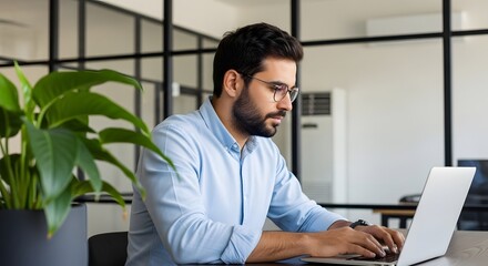 Fototapeta premium Man with glasses and a beard is working on a laptop in an office with a plant and a glass partition