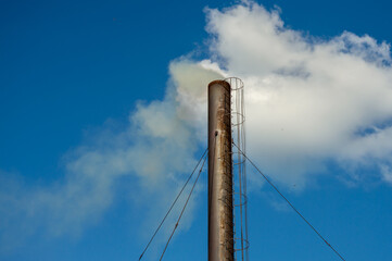 A tall smokestack emits steam into a clear blue sky, framed by fluffy clouds, showcasing an industrial landscape in a city