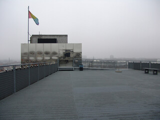Urban rooftop with metal flooring, reflective panels, and a rainbow flag under an overcast sky,...