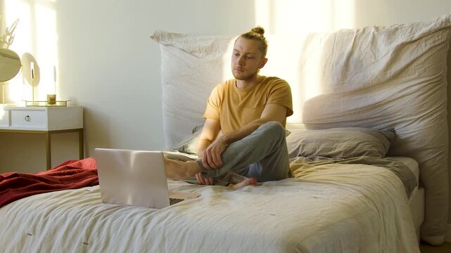 Slow motion 4k 30FPS footage of a Caucasian young man during an online meditation class, he sits in a lotus position in front of laptop monitor in his bedroom.