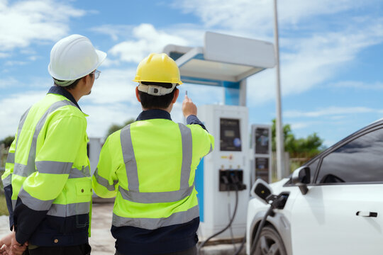 Engineers in safety gear discussing electric vehicle charging station installation at sunny outdoor site - Powered by Adobe