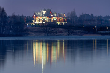 Fototapeta premium Rustic mansion with beautiful lighting and reflection in the water of a frozen river on an autumn evening