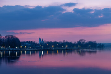Village houses and the church are reflected in the water of the reservoir on a spring evening at sunset