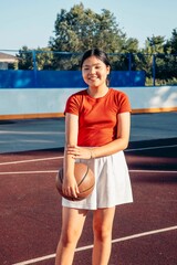 smiling Asian teenage girl in red shirt, white skirt holding basketball on outdoor court on sunny summer day, youth sport girl empowerment healthy lifestyle and leisure physical activity