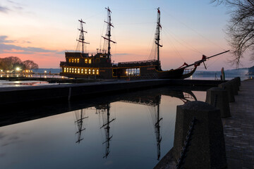 On a spring evening at sunset, an ancient frigate with scarlet sails stands frozen in time at the pier