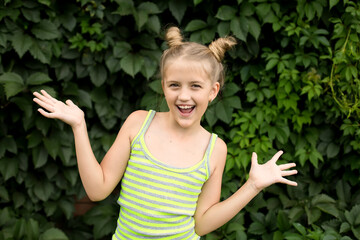 A blonde girl with bunches of hair on her head, standing in front of a leafy hedge, is happy