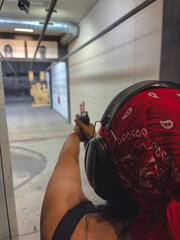 A woman wearing hearing protection and a red bandana is aiming a handgun at paper and silhouette...