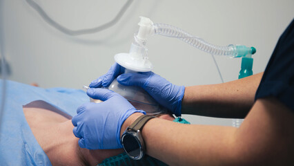 Medical professional in blue gloves administering anesthesia with a bag mask to a patient on an...