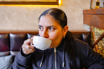 Candid image of a woman enjoying a warm beverage in a cozy indoor cafe setting. Dressed in a dark...