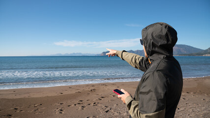 Traveler in a hooded jacket holding a smartphone and pointing toward the ocean horizon while...