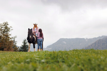 Young woman on horseback, guided by a female western riding school instructor, dressed in plaid...