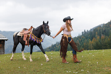 A young woman in a cowboy hat and chaps walks a black and white horse with a saddle across a grassy...