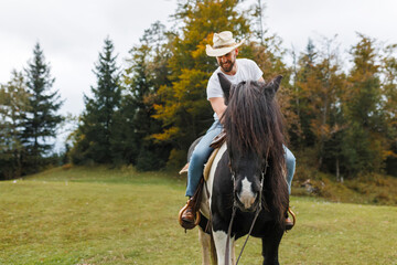 A man wearing a cowboy hat rides a black and white horse as a woman in a plaid shirt smiles and speaks to him in a grassy field.