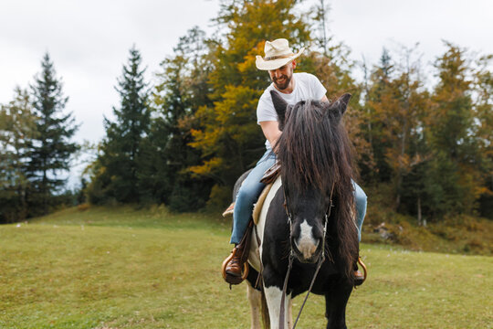 A man wearing a cowboy hat rides a black and white horse as a woman in a plaid shirt smiles and speaks to him in a grassy field. - Powered by Adobe