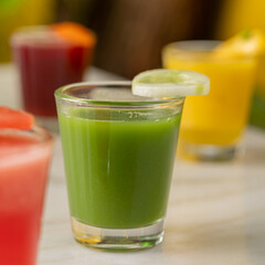 a close-up of a vibrant green health shot served in a clear glass, garnished with a fresh cucumber slice. In the blurred background, colorful juice shots in red, yellow, and orange 
