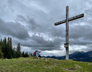 Zwei Wanderer sitzen bei bewölktem Himmel am Gipfelkreuz des Sulzberg und einer deutet ins Tal,...
