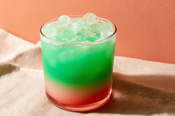 A refreshing layered cocktail with green, white, and red hues served on ice in a clear glass, set against a pink wall and beige fabric background