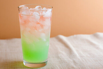 A refreshing glass of iced drink with a gradient from green at the bottom to clear at the top, sitting on a light fabric surface against a peach background