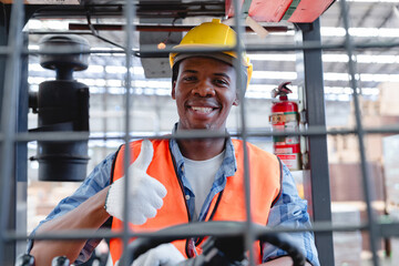 Inside view of African forklift driver wearing yellow helmet and orange vest, posing with thumbs up...