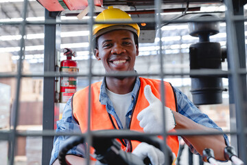 African male forklift operator wearing safety gear smiles and gives thumbs up while working in...
