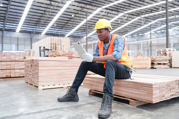 African male worker sitting on stacked wood pallets inside warehouse, wearing safety helmet and vest, checking data on digital tablet. Modern pallet factory setting shows use of smart technology