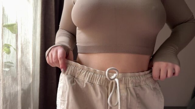 A young woman smiles and proudly shows her dramatic weight loss by wearing old jeans that are now too big. She celebrates her fitness success at home.