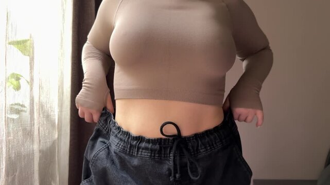 A young woman smiles and proudly shows her dramatic weight loss by wearing old jeans that are now too big. She celebrates her fitness success at home.