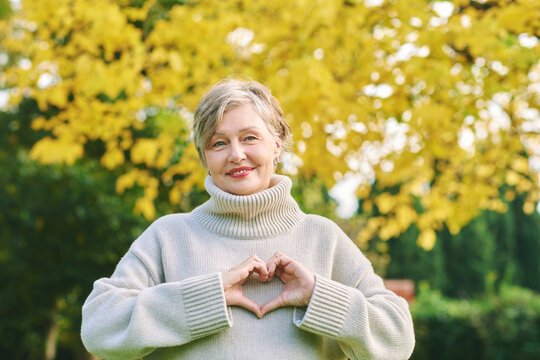 Close up portrait of happy mature 55 - 60 year old woman posing in autumn park, active and healthy lifestyle. Lady showing heart shaped hand sign, love and gratitude