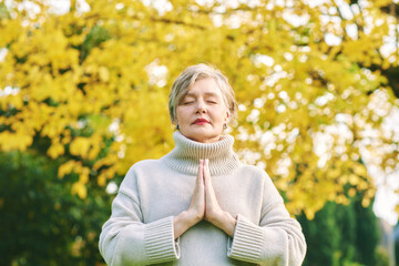 Outdoor portrait of mature woman meditating in autumn park, showing Anjali mudra, honoring and celebrating this moment