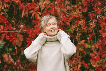 Autumn portrait of happy mature woman wearing beige pullover or sweater, posing outdoors with red ivy red leaves background