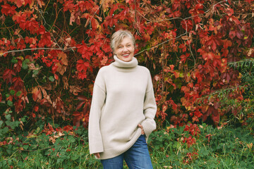 Autumn portrait of happy mature woman wearing beige pullover or sweater, posing outdoors with red ivy red leaves background