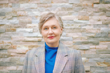 Portrait of 55 - 60 year old woman with grey hair, wearing jacket, posing on brick background