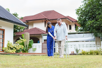 Home caregiver assists elderly man with walker in garden, providing support and encouragement in...