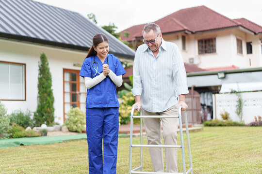 Home caregiver supports elderly man using walker in garden, cheerful atmosphere, outdoor rehabilitation, residential care, health recovery, sunny day, encouragement and compassion